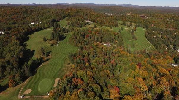 Aerial view of a golf course surrounded by fall foliage-covered hills under a clear sky.