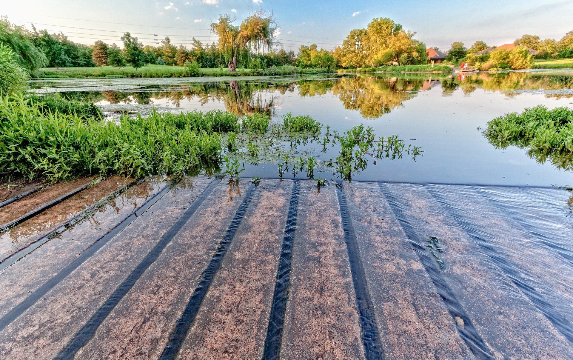 Water flows over a series of dark, textured spillway steps into a calm pond surrounded by trees on a sunny day.