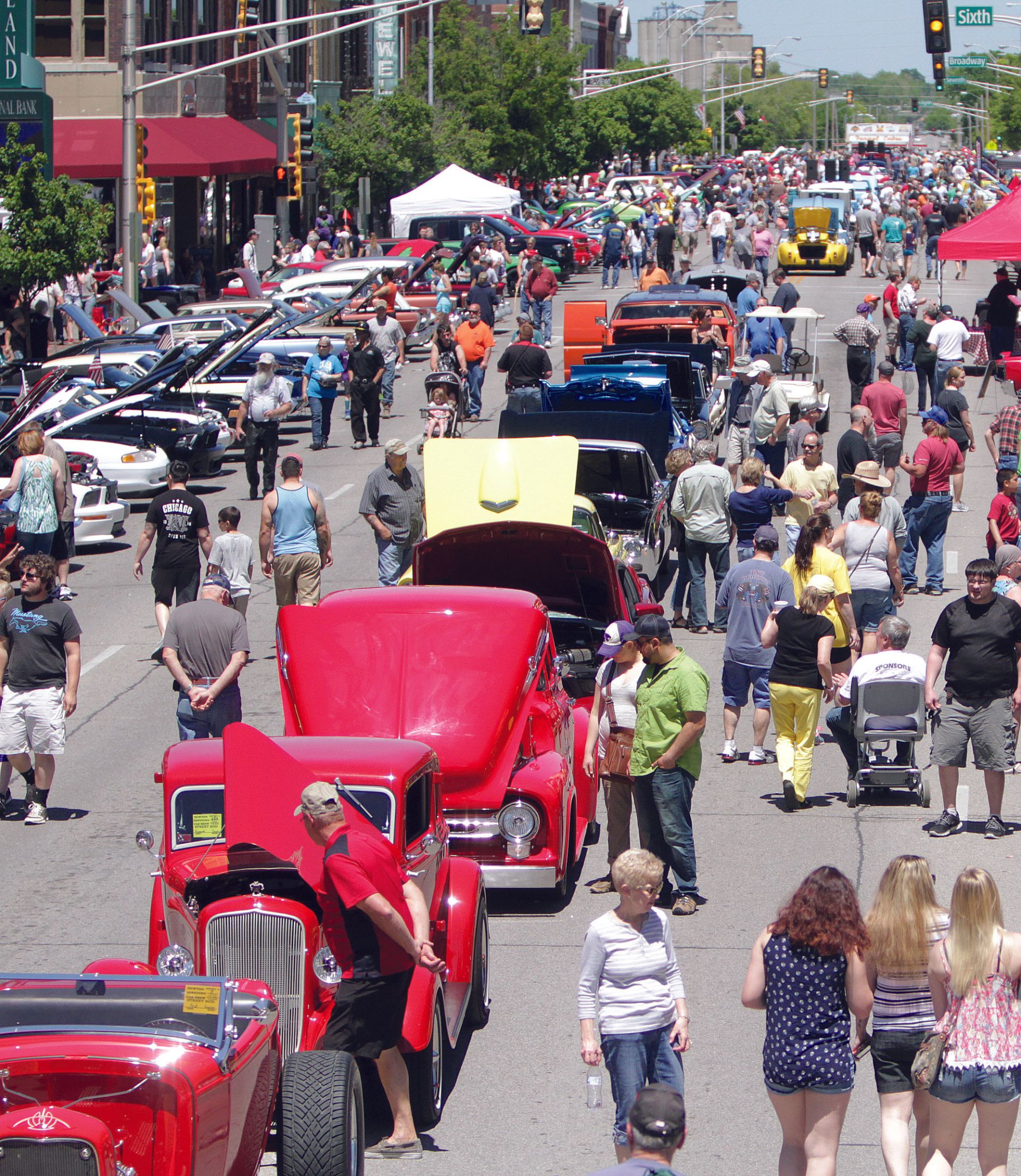 A street fair featuring classic cars on display with their hoods open, surrounded by pedestrians on a sunny day.