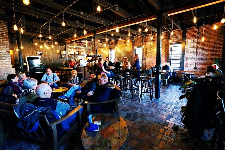 A rustic, industrial-style room with brick walls, exposed beams, and hanging lights where people socialize at tables.