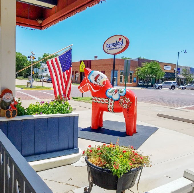 A large red Dala horse statue stands on a street corner next to an American flag and a blue planter with flowers.