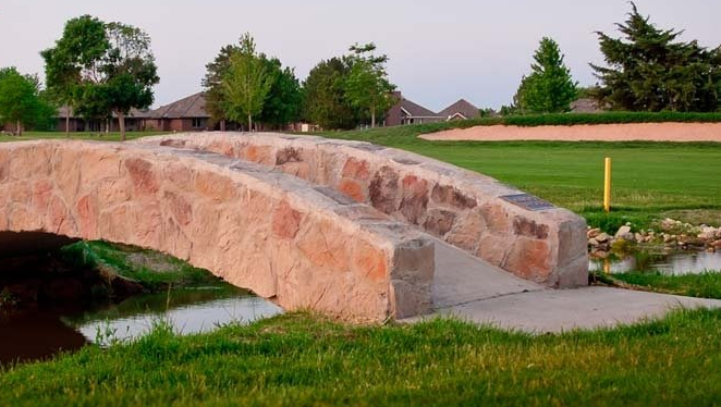 A stone arched footbridge spans a small stream on a green golf course near a sand bunker and houses.