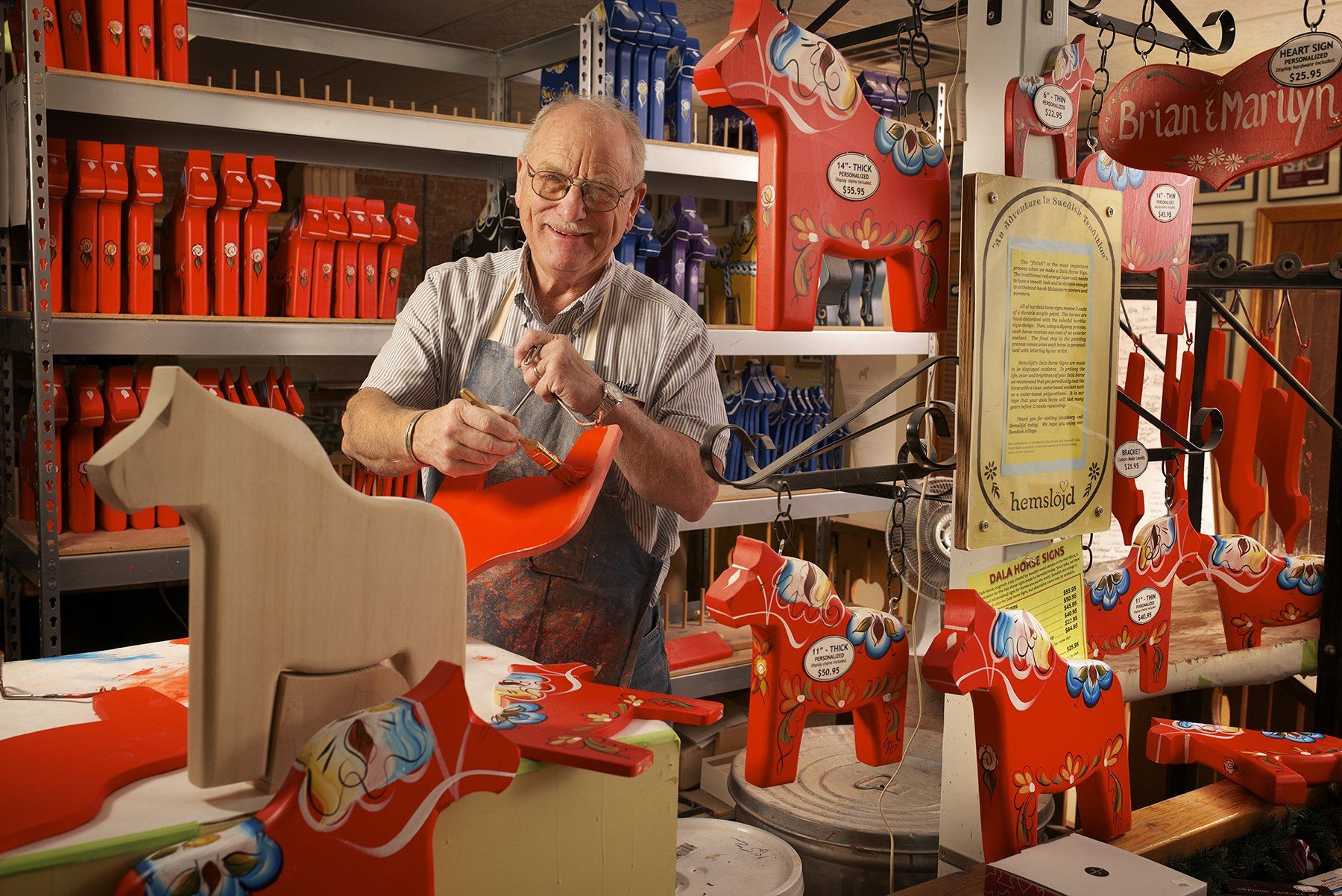 A person in a workshop painting a wooden Dala horse, surrounded by shelves stocked with finished red Dala horses.
