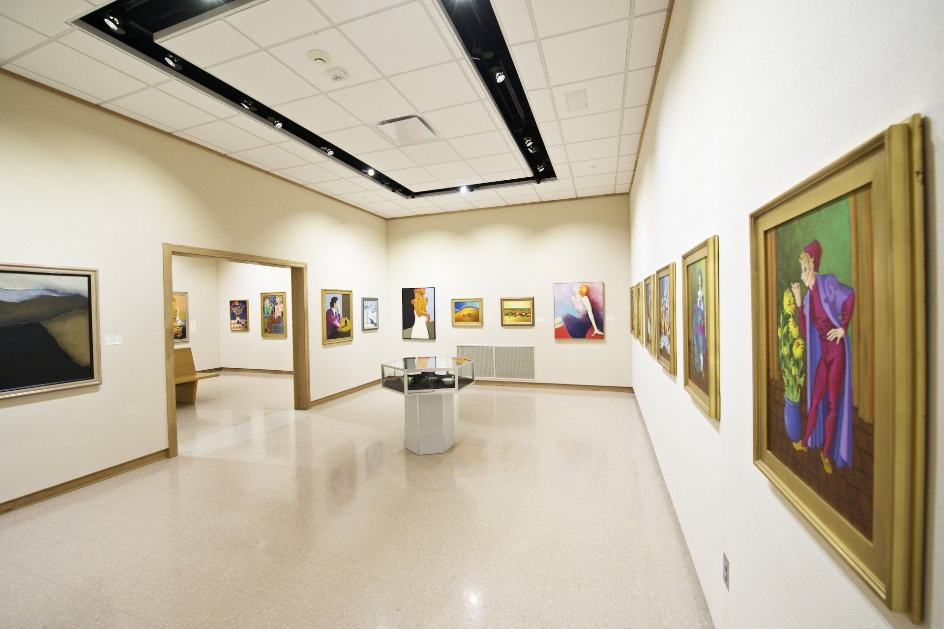 A bright art gallery with paintings on white walls, a central display case, and a doorway to another exhibition space.