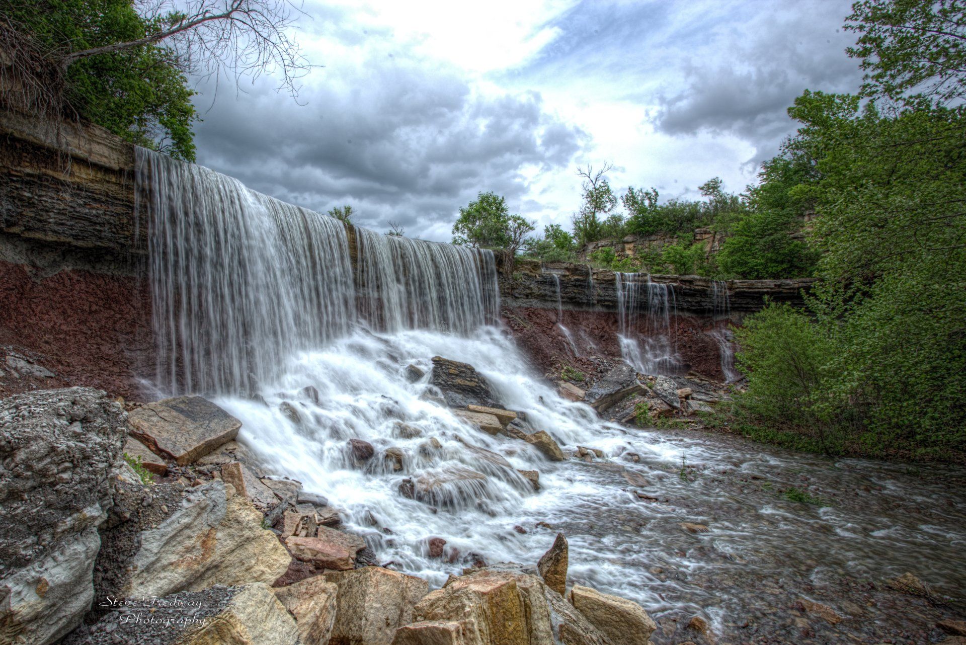 A waterfall cascades over a layered, reddish-brown rock cliff into a rocky stream under a cloudy sky.