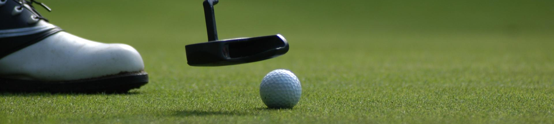 A golf club putter is positioned over a white golf ball on a green putting surface, with a golf shoe visible to the left.