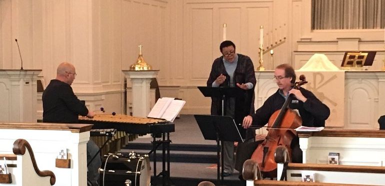 Musicians in a church: a cellist, a man at a keyboard, and a speaker at a podium.
