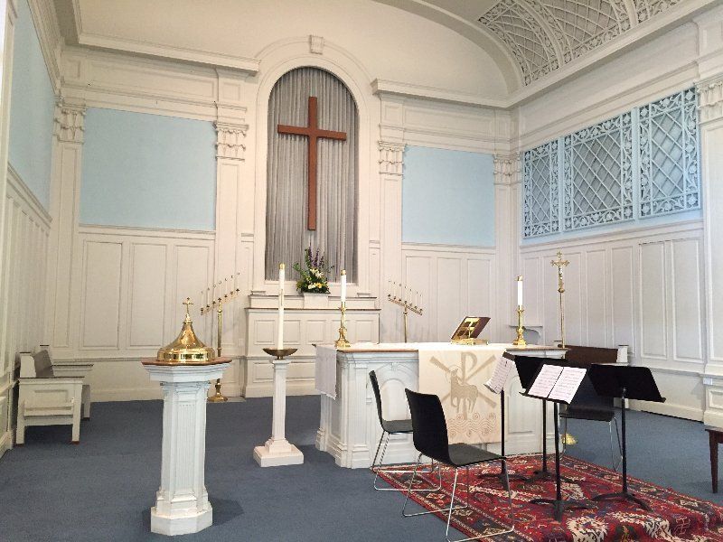 Interior of a church. White paneled walls, a cross above the altar, candles, and chairs on a blue carpet.