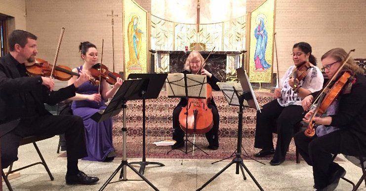 String ensemble performing in a church with stained glass. Musicians seated, playing instruments: violin, cello.