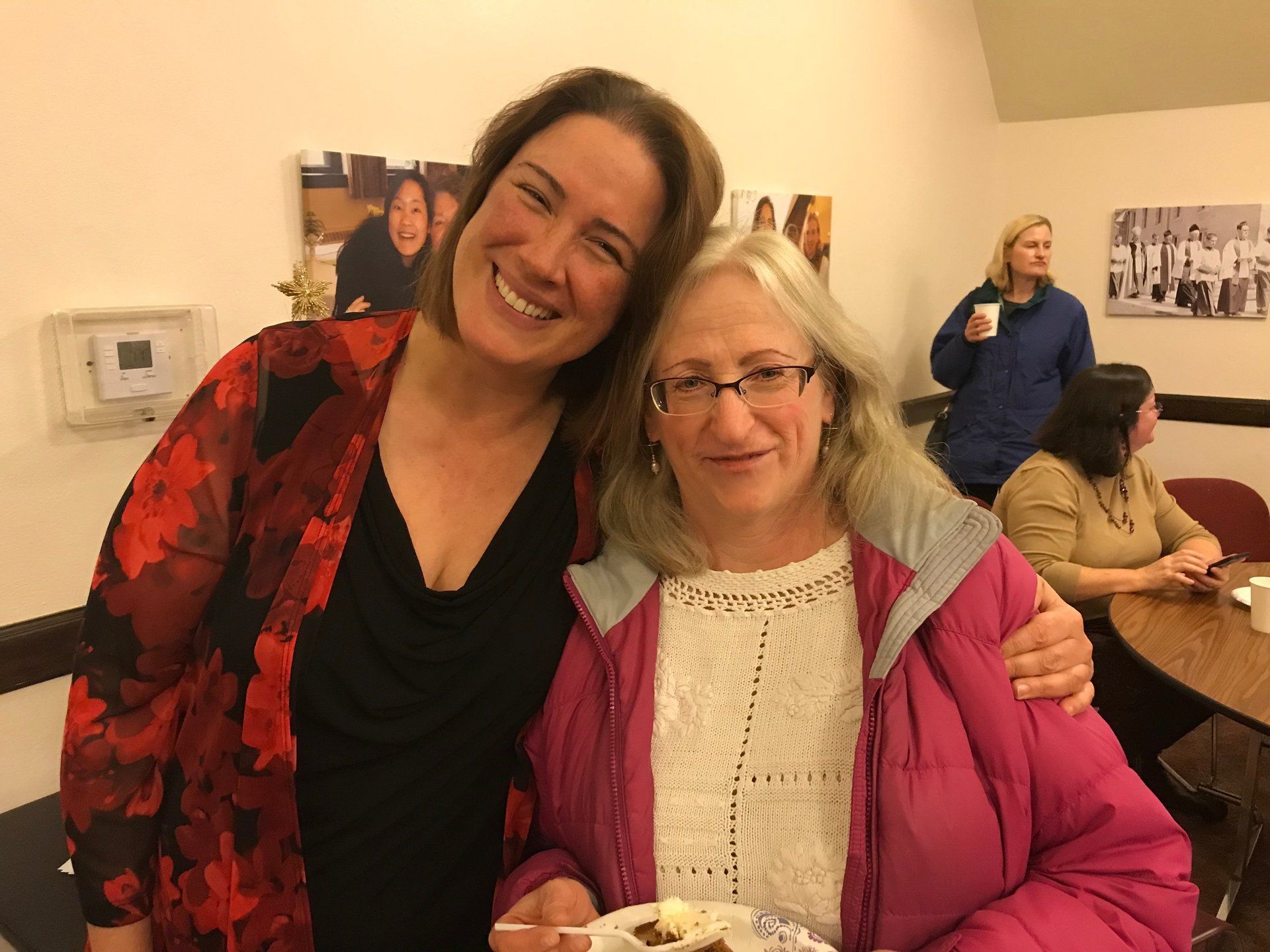 Two women smiling, arm in arm, indoors. One in a floral jacket, the other in a pink coat. Others in background.