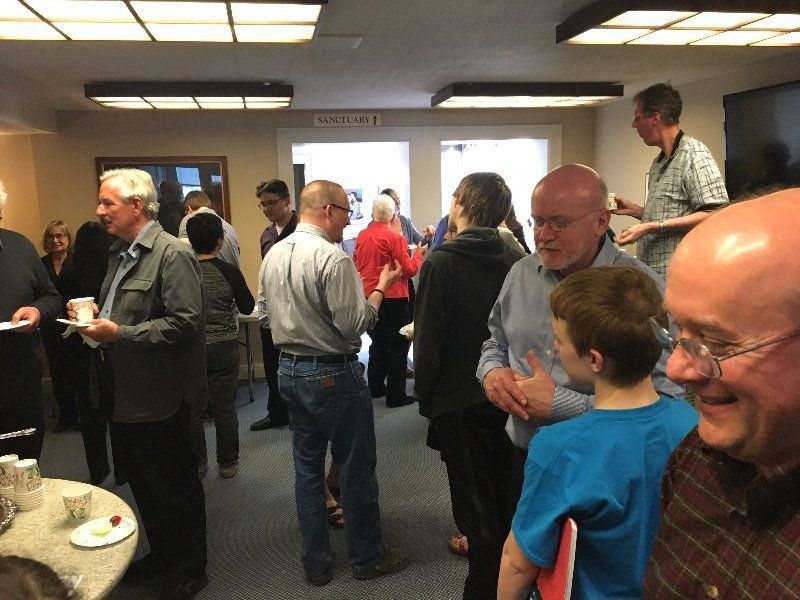 People socializing in a room, some holding food/drinks. Various clothing styles, neutral lighting, indoor setting.