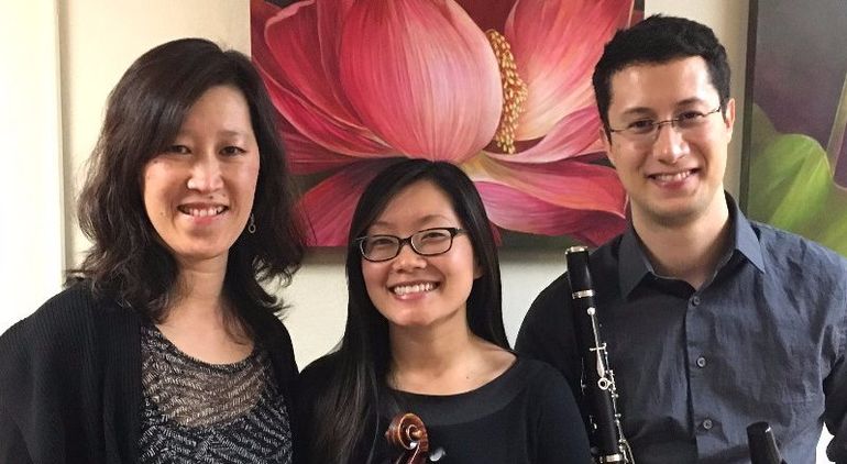 Three smiling musicians stand in front of a flower painting. The person on the right is holding a clarinet.