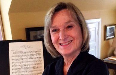 Woman smiles, stands near sheet music, in a room with warm-toned walls.