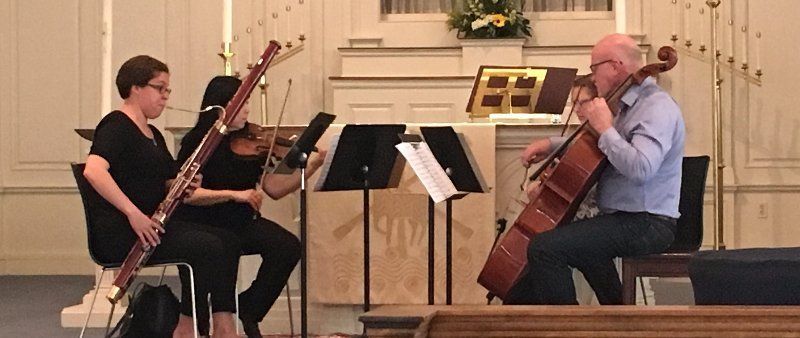 Three musicians playing instruments on a stage. One with a bassoon, one with a violin, and one with a cello.