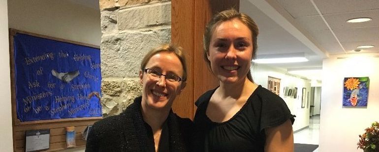 Two smiling people stand in a hallway with art on the walls. One wears glasses and a black jacket. The other wears a black shirt.