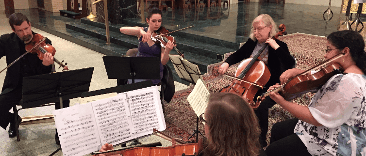 String quartet performing in an interior space. Musicians are playing violins and cello, using sheet music on stands.