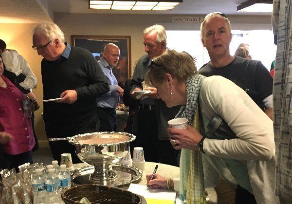 People at a buffet table, one writing, others serving food from a large bowl, indoors.
