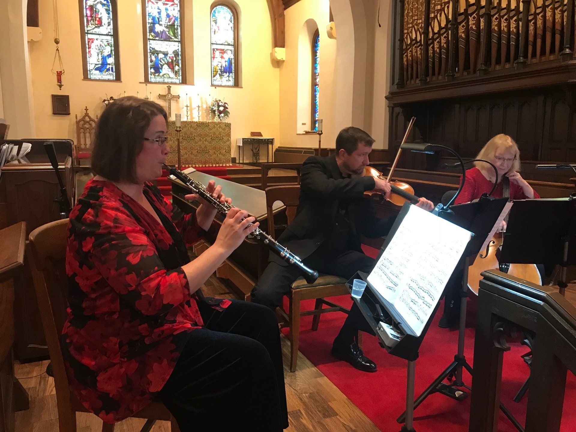 Three musicians performing in a church: oboe, violin, and cello, with sheet music, red accents, and stained glass.