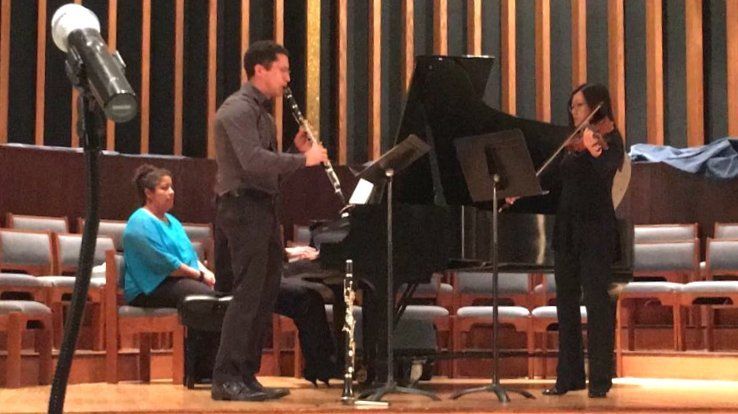 Oboe player, violinist, and pianist performing on stage, with an audience member.