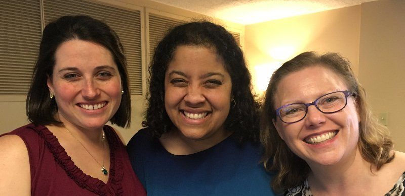 Three smiling women posing closely together indoors, lit by warm light.