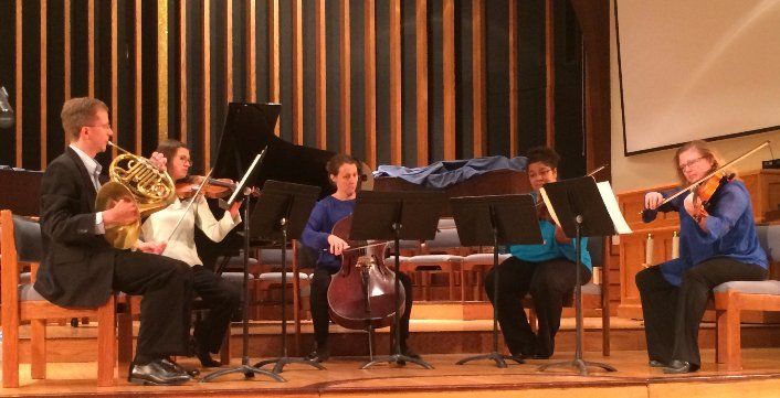 A musical ensemble performs in a wood-paneled room. Musicians play various instruments including violin and cello.