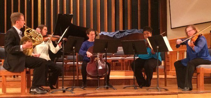 A musical ensemble performs in a wood-paneled room. Instruments include a French horn, violin, cello, and piano.