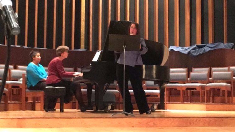 Woman singing, pianist at piano, another woman seated. Performance on stage in auditorium.
