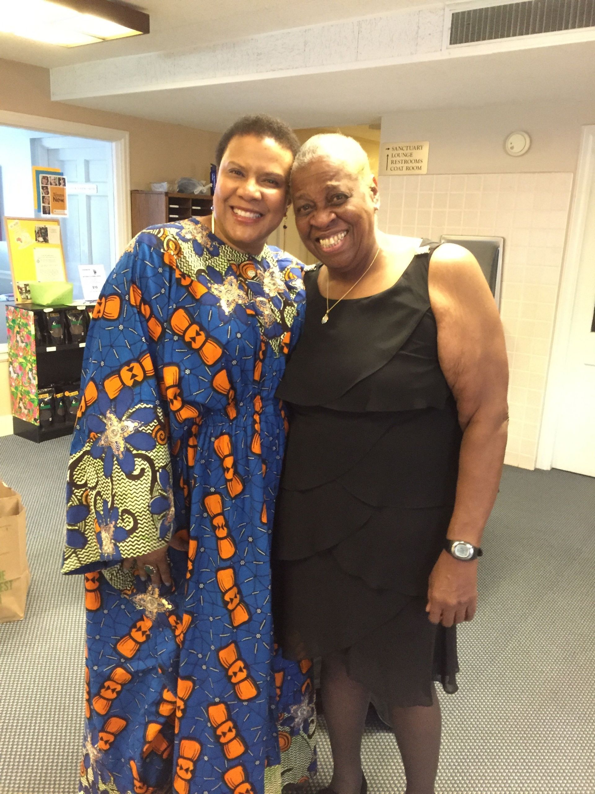 Two women smiling and posing indoors. One wears a blue patterned dress, the other a black dress.