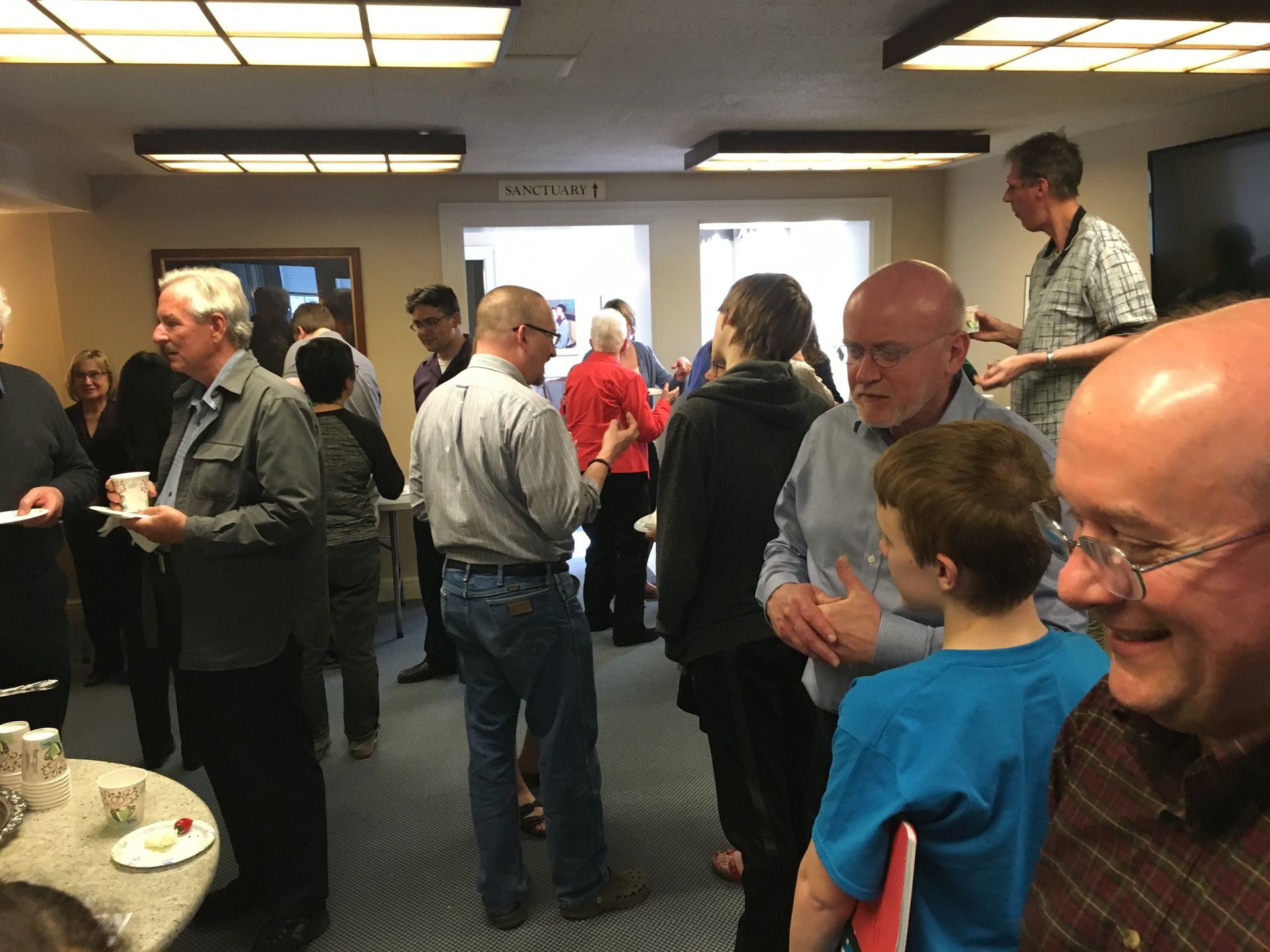 People socializing indoors, some holding food and drinks. Brightly lit room, casual attire, multiple conversations.