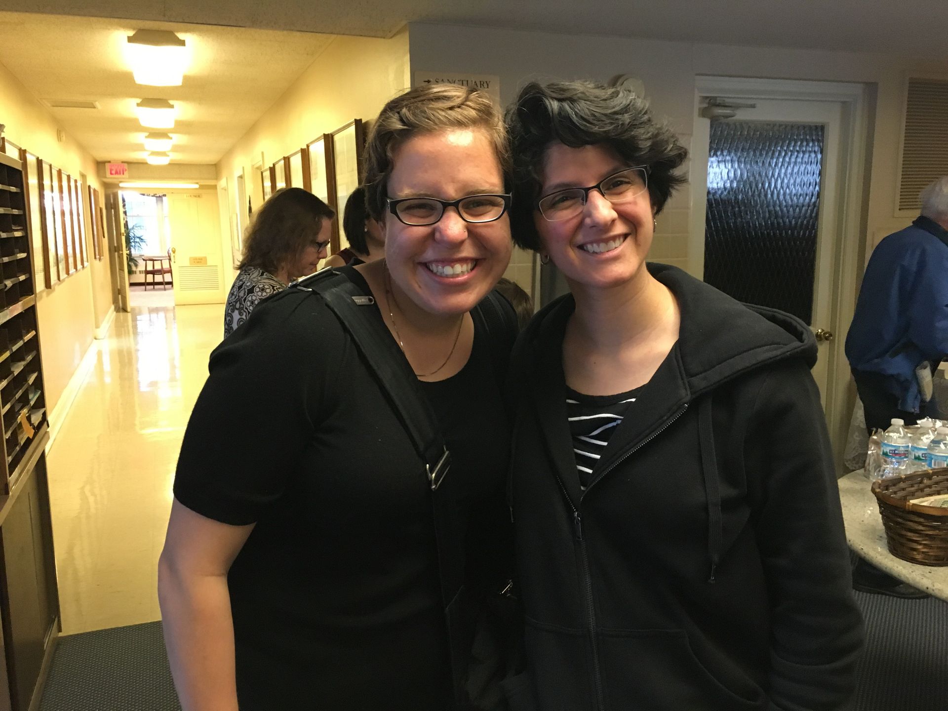 Two women smiling, posing in a hallway with art on the wall and a doorway at the end.