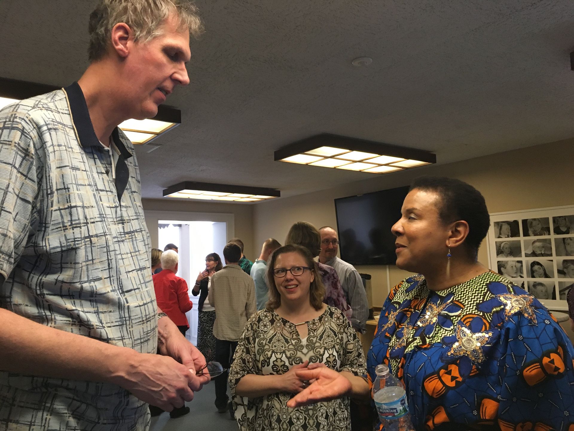Man and woman conversing, others in background, indoors. Woman wears patterned blue top, man in printed shirt.