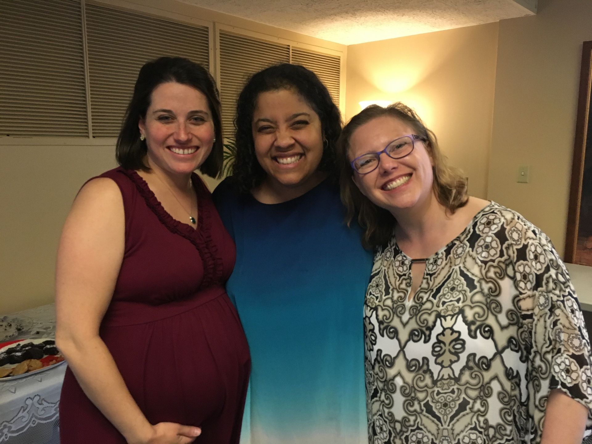 Three smiling people, one pregnant, stand close together indoors. They are lit by warm light and appear to be at an event.