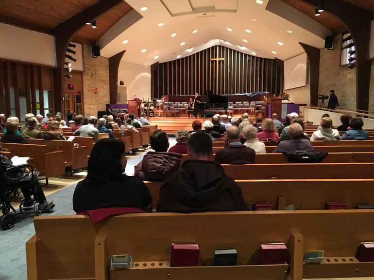 Audience in a church watches a person perform on stage with a piano and cross backdrop.