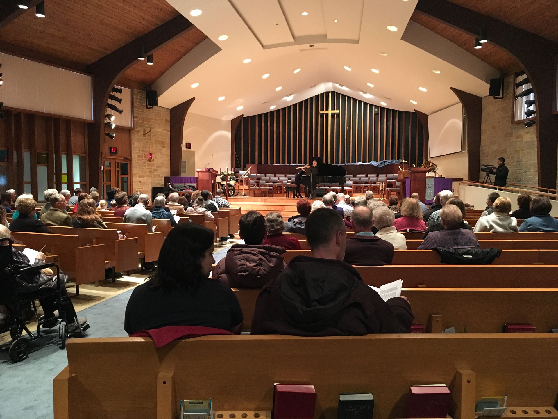 People seated in a church sanctuary, facing a stage with a cross and a piano.