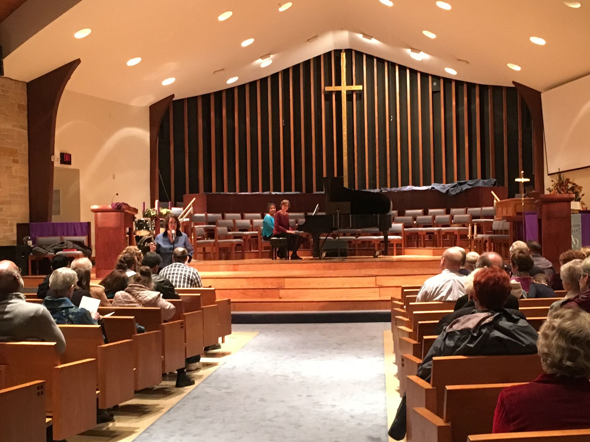 People seated in a church auditorium, listening to a piano performance on stage with a cross.