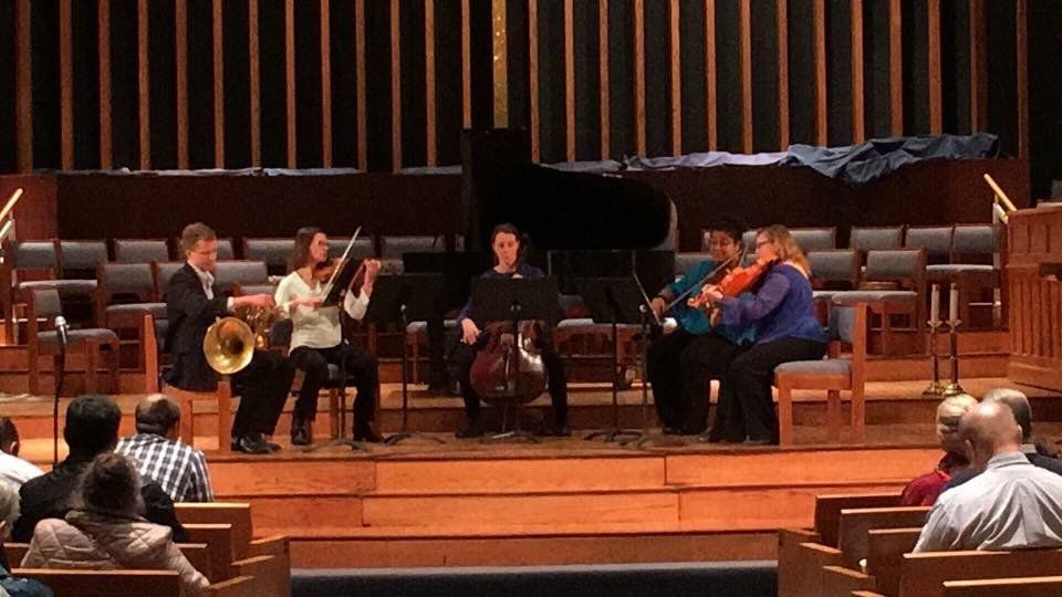A musical ensemble performing on stage with an audience. Musicians play violin, cello, and other instruments.