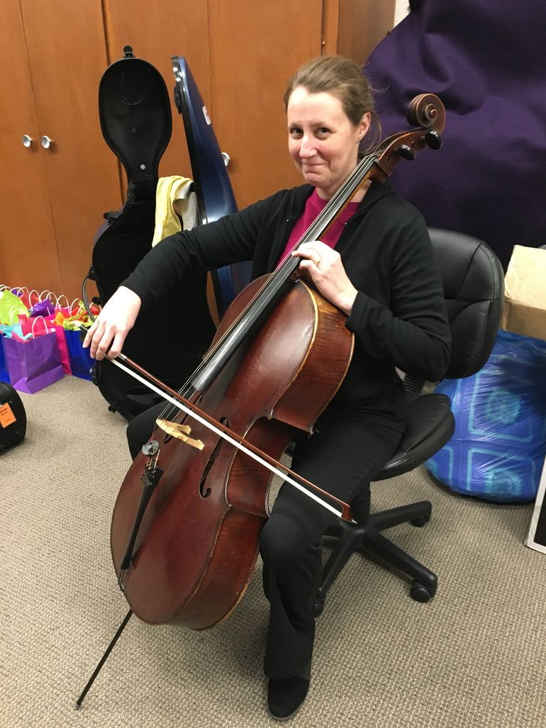 Woman seated, holding a cello and bow. She's smiling, in a room with a closed case and storage behind her.