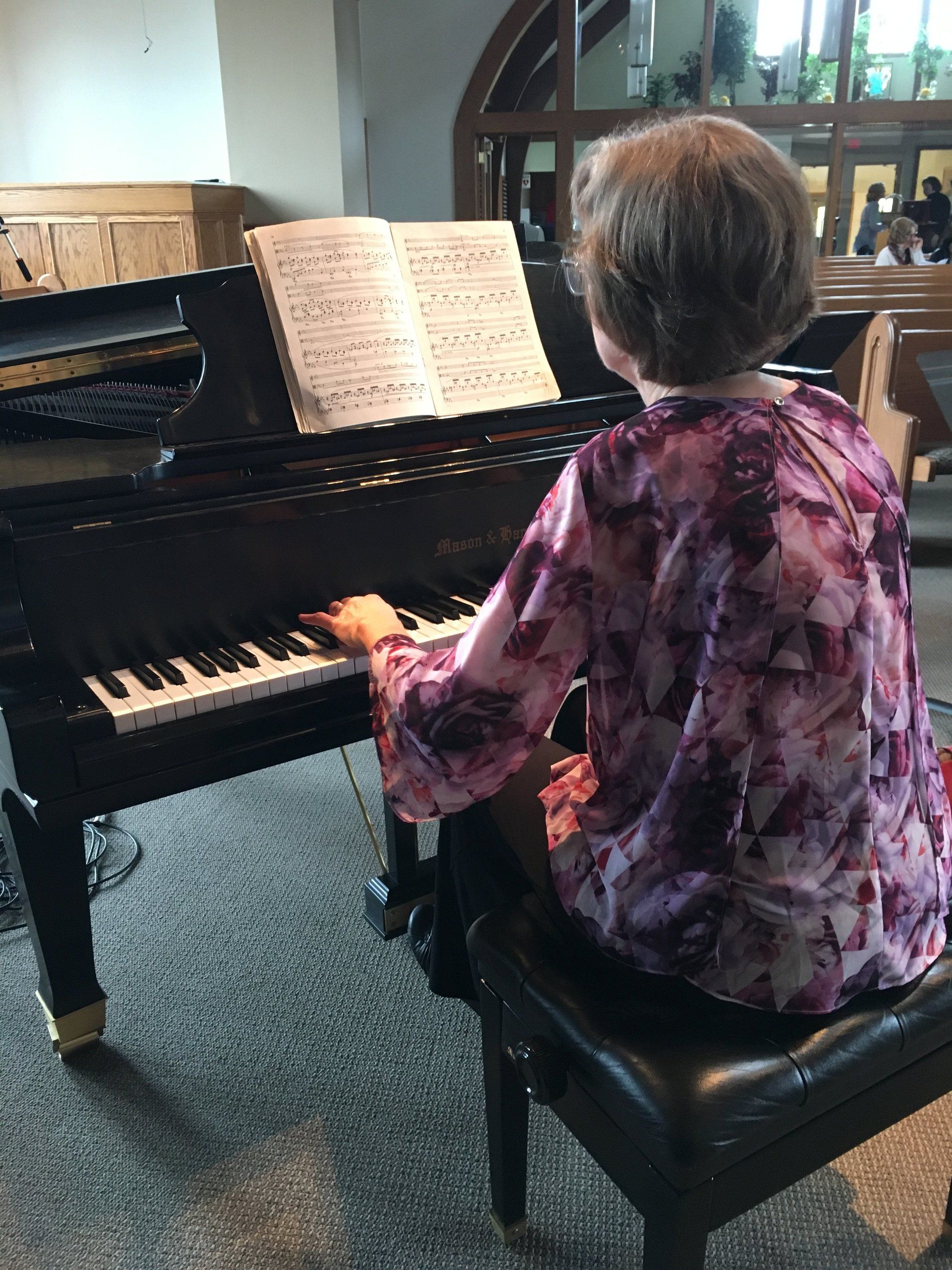 Person playing a grand piano, reading sheet music. Wearing a purple floral top, seated on a piano bench. Indoors.