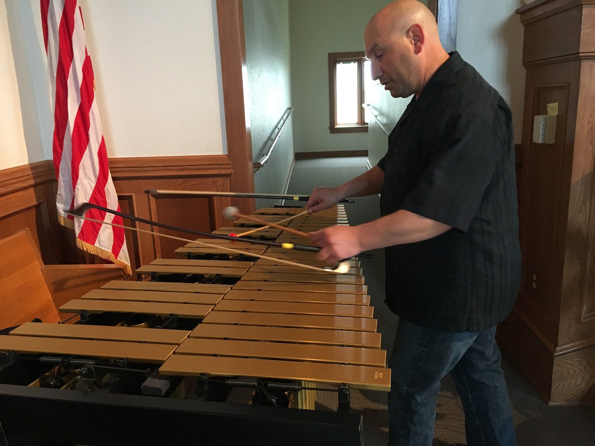 Man playing a marimba indoors, with an American flag to the left and stairs in the background.