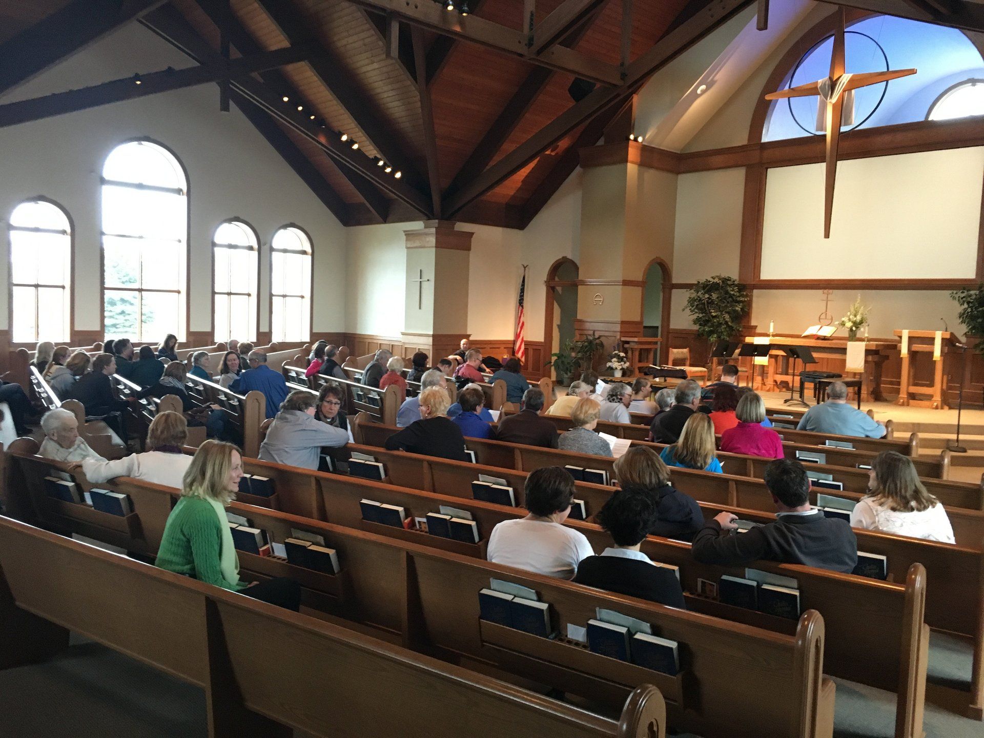 People seated in pews during a church service; wooden ceiling, cross, and altar visible.