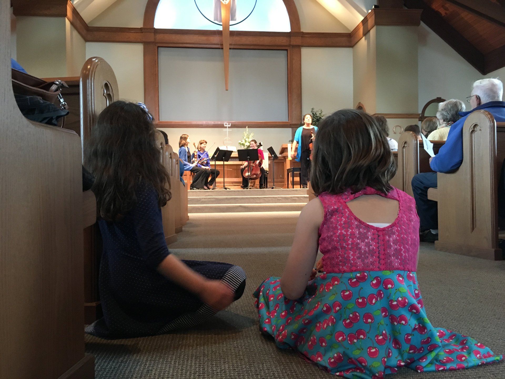 Two girls sit on the floor of a church, watching musicians on the stage.
