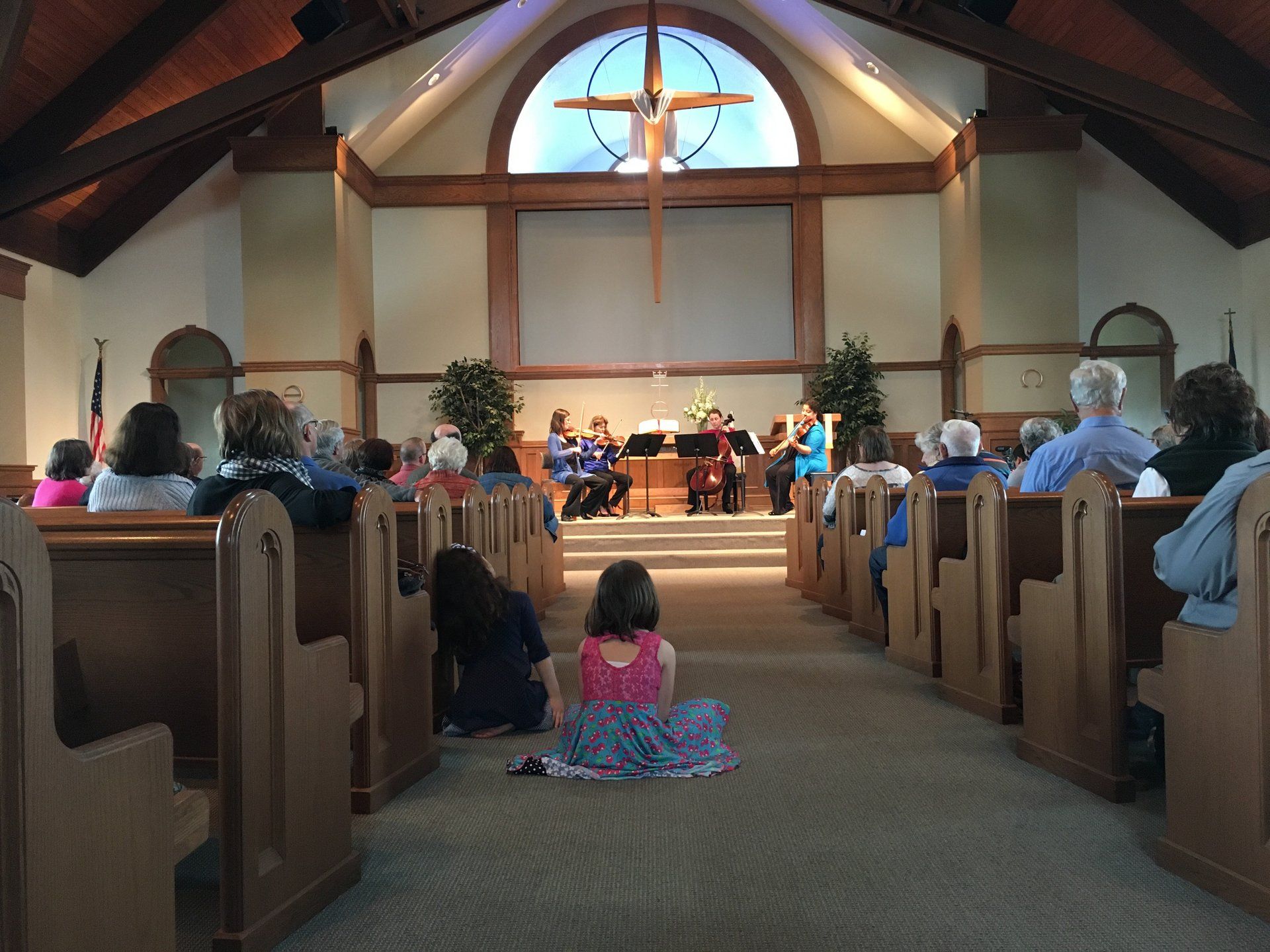 Interior church view, string quartet playing, audience seated in pews, cross above stage.