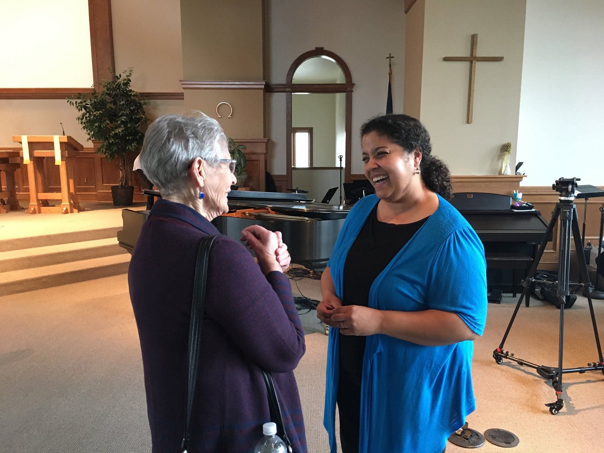 Two women converse and laugh inside a church. One wears a blue sweater, the other a purple jacket. A wooden cross hangs on the wall.