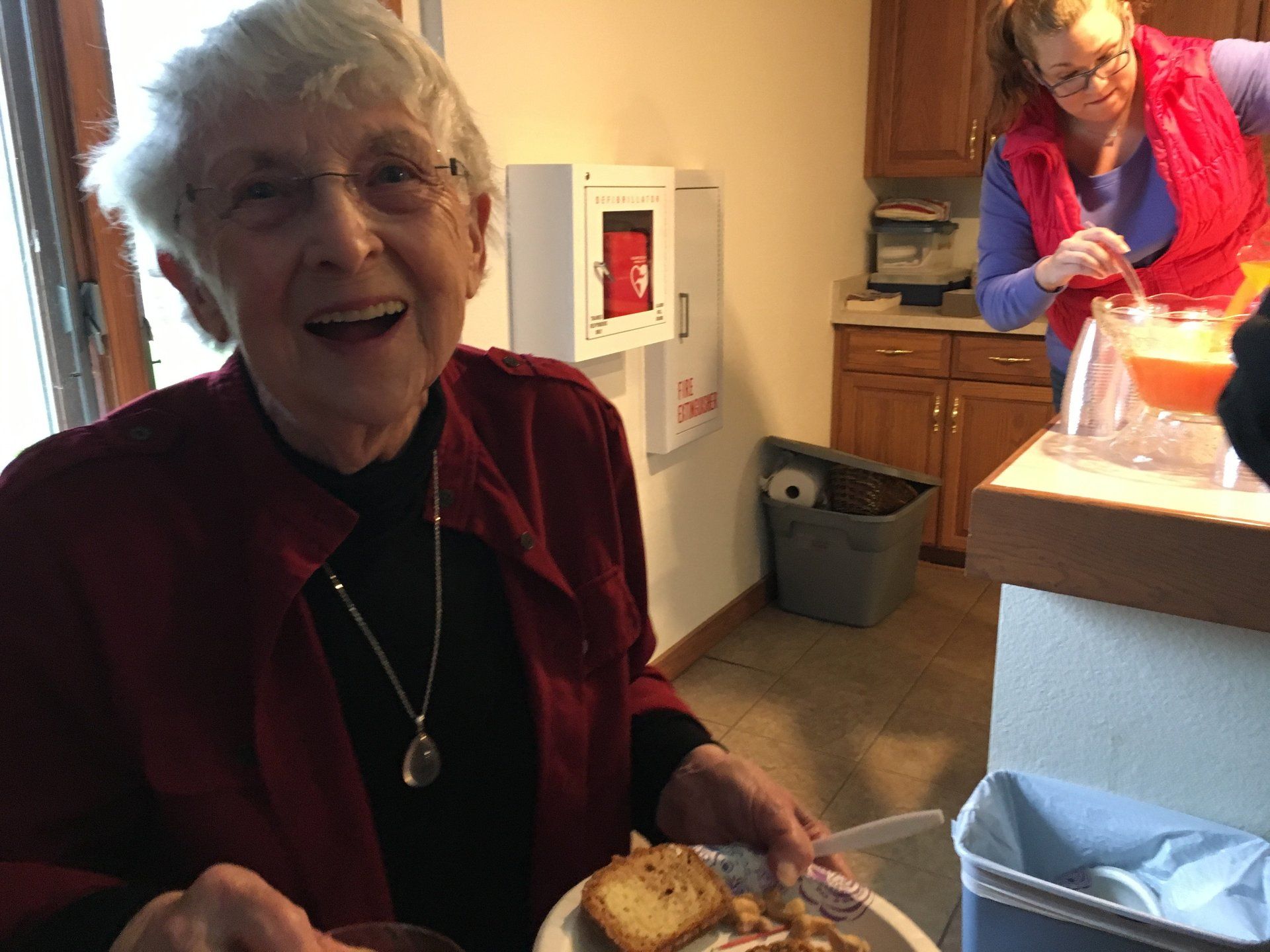 Woman smiles, holding a plate of food. Another woman in background, near a kitchen counter.