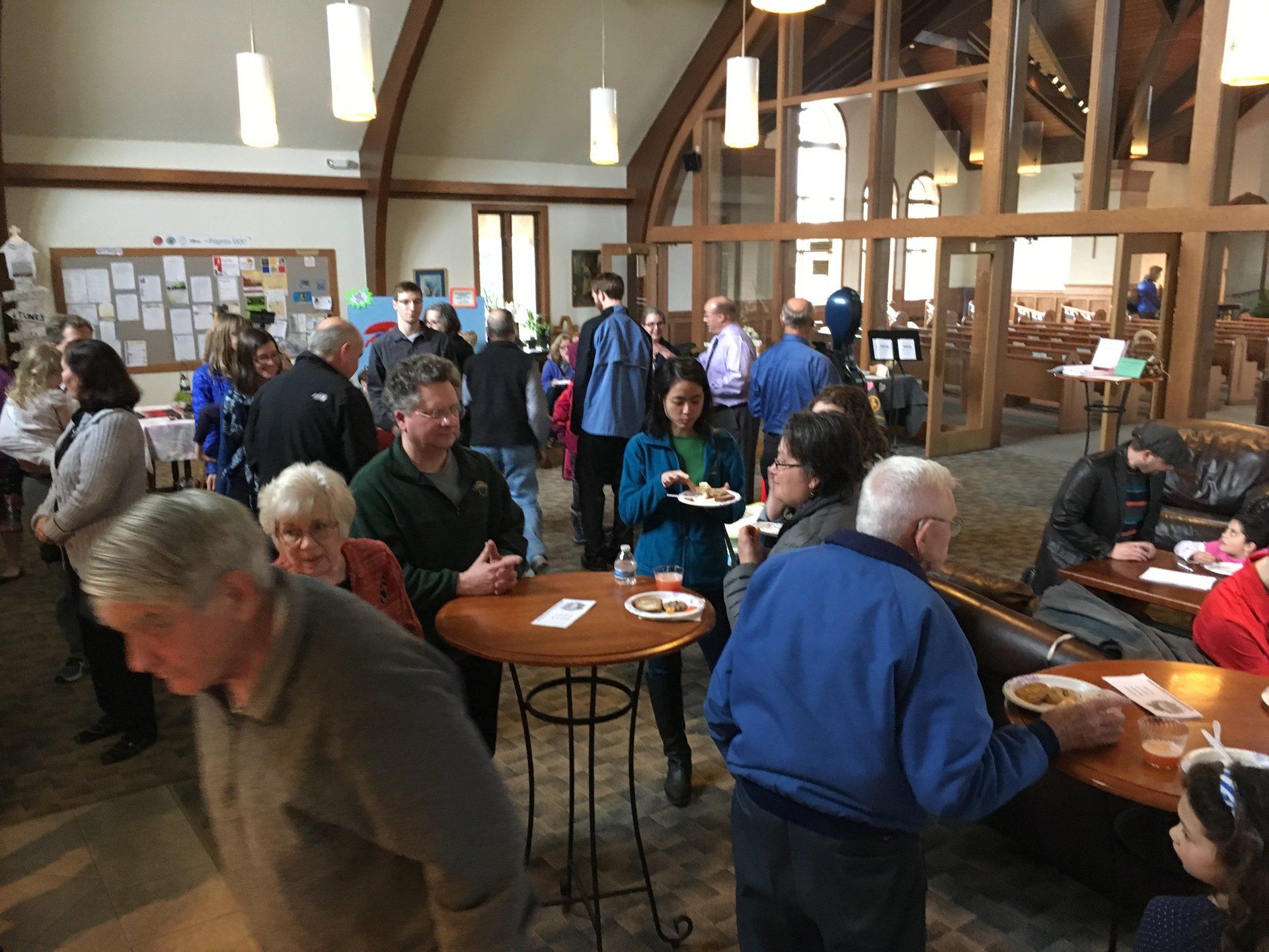People socializing and eating at an event inside a building with large windows and wooden beams.