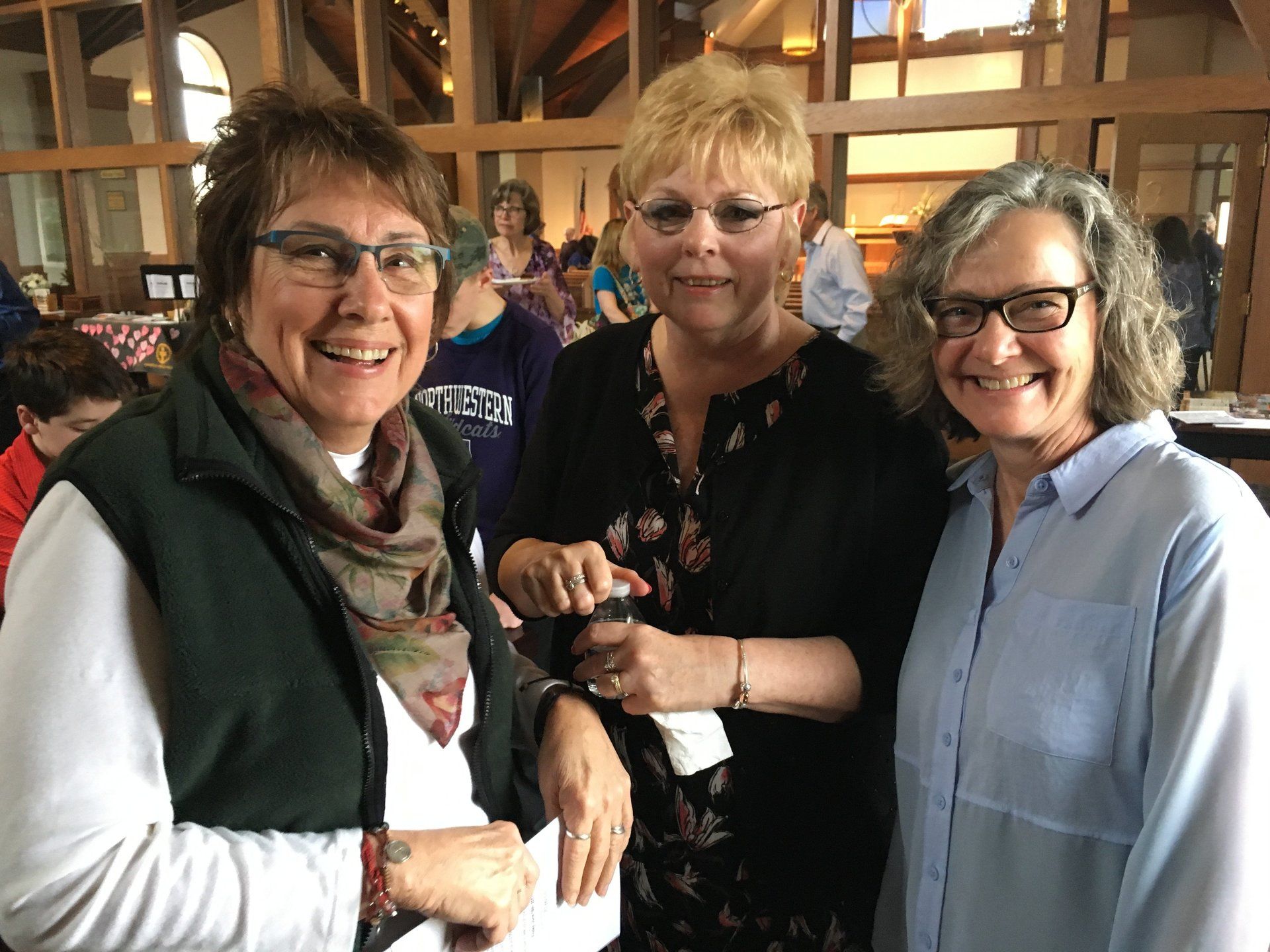 Three women smiling indoors, one wearing a green vest, one with a floral top, and the third in a blue shirt.