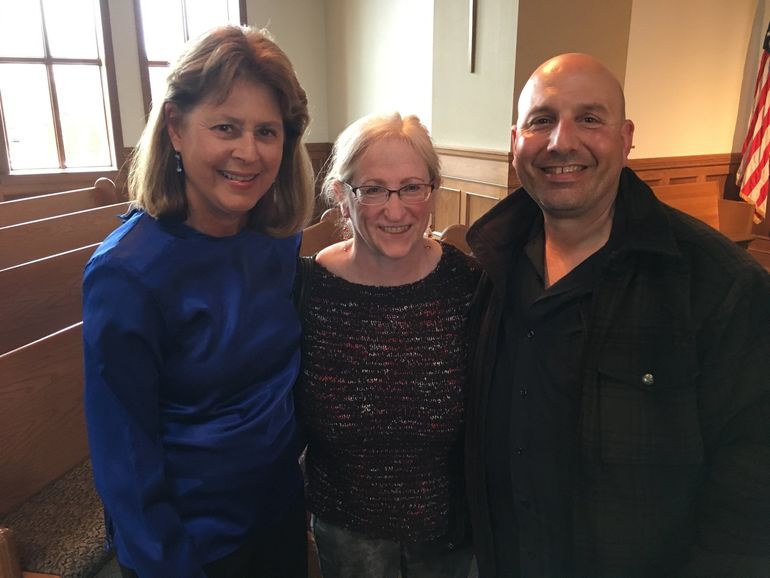 Three people smiling, posed inside a building, one wearing blue, center woman in glasses, right man in a black jacket.