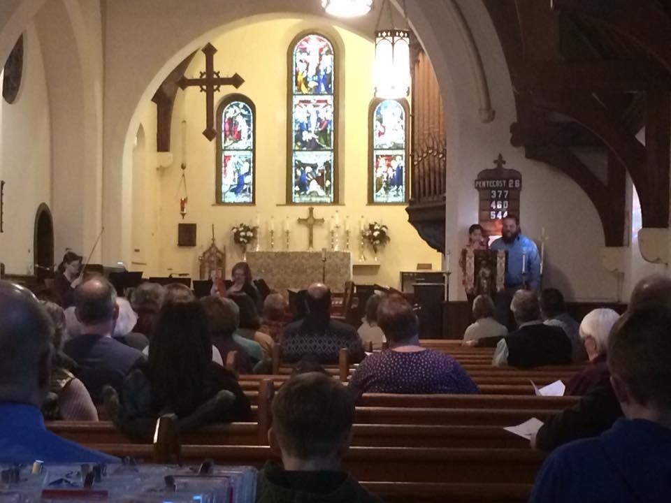 People in church pews watch a presentation. Two people stand near an organ and stained glass windows, in a church.