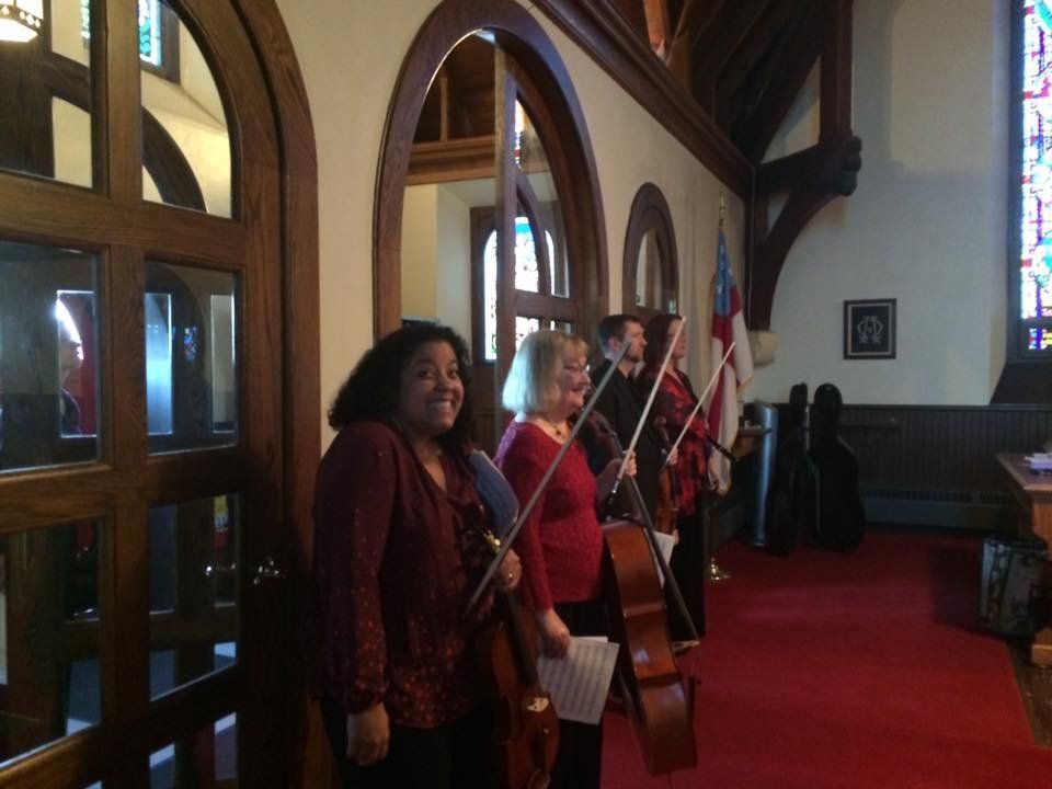 Musicians with string instruments inside a church, standing on a red carpet.