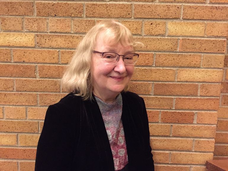 Woman with glasses smiles in front of a brick wall. She wears a black blazer and patterned scarf.